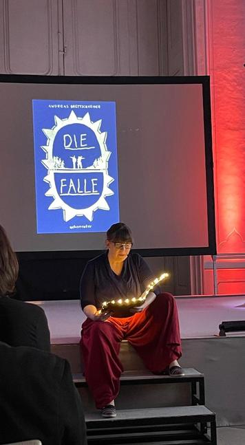 Ich sitze auf der Treppe der Bühne und lese aus "Die Falle" von Andreas Brettschneider. Mit meiner coolen, leuchtenden Mappe. 🥰