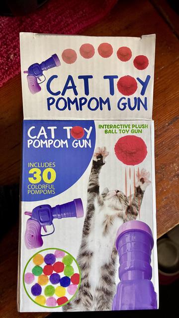 On a redwood countertop, a box with a pink and purple toy gun that looks like it belongs in Looney Tunes with a bunch of colorful pompoms and a young grey tabby cat jumping with arms outstretched.