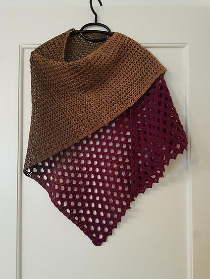 A crocheted shawl is hanging on a black hanger against a white door. The shawl has a gradient color transition from brown at the top to a deep burgundy at the bottom. The upper part of the shawl has a tighter, more solid stitch pattern, while the lower part features a more open, lacy stitch pattern with openwork holes.