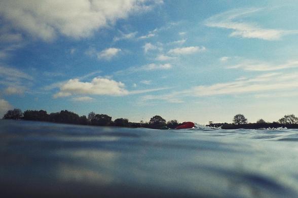 A horizontal colour photograph of a swimmer's arm mid stroke in the blue of a lake. The foreground is all water. In the background are some trees.