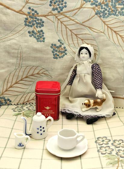 Tea tableau in pale colors featuring a small bisque doll in a pinafore and bonnet holding a puppy on her lap. She is about to partake of tea from a tiny tea set. She is ensconced on and in front of beautiful embroidered linen fabrics. The tea is English Afternoon black loose-leaf.
