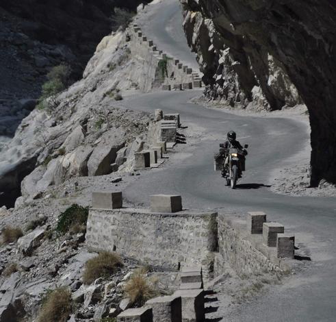 Ein Motorrad auf einer kurvigen grauen Straße die rechts von Felswänden und links von einem Abgrund begrenzt wird