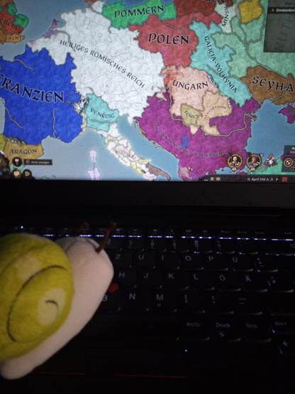 Eine gelbgrüne, süße Kuscheltierschnecke sitzt links auf einer beleuchteten Thinkpadtastatur und kuckt auf den Bildschirm, auf dem eine Europakarte im Spiel Crusader Kings III zu sehen ist.