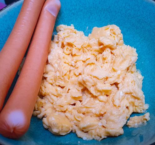 In einer Bowl liegen zwei Wiener Würstchen und eine Portion Rührei aus sechs Eiern. Das Rührei ist goldgelb und in kleine Stücke zerteilt. Die Würstchen sind nebeneinander gelegt. Die Bowl in der alles liegt hat eine glatte Oberfläche und ist kräftig türkisfarben.