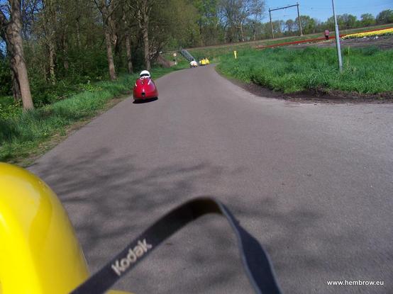 Colourful velomobiles going past a colourful field of  flowers. With a cameo from my camera strap.