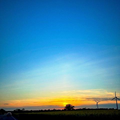 A scenic view of a sunset with vibrant orange and blue hues in the sky. Wind turbines are visible on the right, and a tree silhouette is present near the horizon. A car is driving along a road in the foreground.
