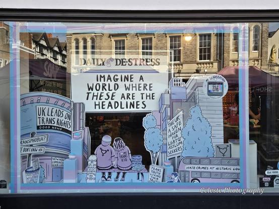 the window display of the store

it is completely dedicated to the display, everything trans colours, it depicts a street scene with drawn characters and some fake headlines showing positive news, including "GPs on board with gender-affirming care", "unrealistic wait times binned", "UK leads on trans rights", "UK safe haven for LGBTQ asylum seekers", "Hate crimes at historic low"

at the top is the problem: "IMAGINE A WORLD WHERE THESE ARE THE HEADLINES"