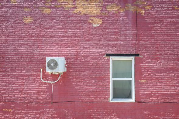 A white window with a black horizontal bar over it and a small air conditioner in/on a brick building painted pink.  Some of the pink paint is peeling off revealing the brick underneath the paint.