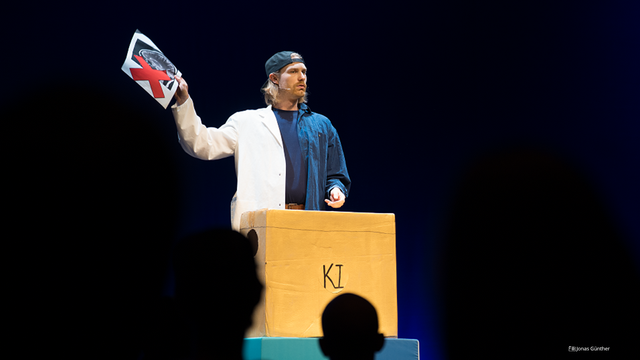 Foto von Marco Gustav bei seinem FameLab-Aufritt. Er steht vor einem Karton, auf dem KI geschrieben steht und hält in seiner rechten Hand ein Blatt Papier.