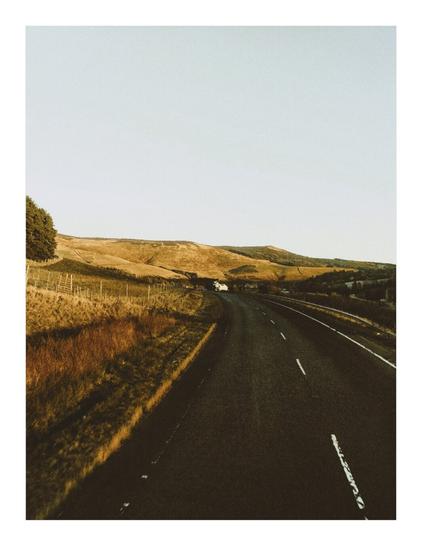 A vintage style photo of a country road. On the left there are grassy fields which range in colour from a dark yellow, orange and a deep dark green. The road is black tarmac and stretches forward to the middle of the photo and turns off towards the left into the distance. There is a white building in the middle of the photo. The top half is a bright sky with no clouds.