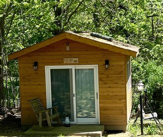 Photo of a 12-foot by 12-foot rustic wooden office surrounded by trees with spring-green leaves. It has a sliding glass door and a small porch with a rocking chair.