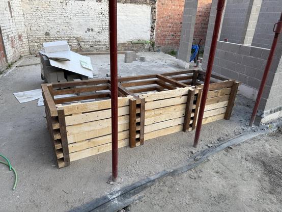 The image features a construction site with wooden pallets arranged in a rectangular formation, likely for composting. The background includes scattered construction materials and partially built walls.