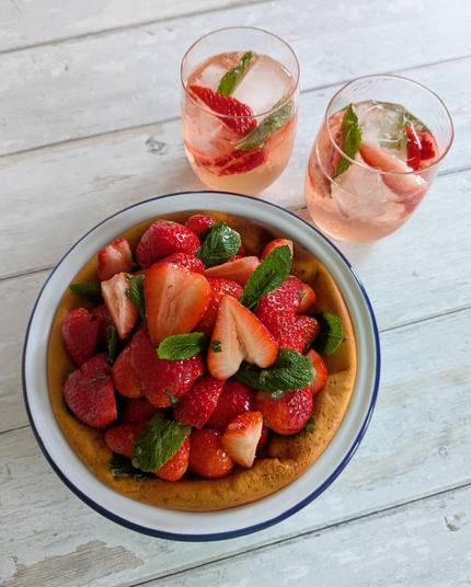 A rectangular photo of a pie and two cocktails sitting on a white wooden background. The pie is a golden-brown colour below a huge pile of bright red strawberries, with green mint leaves poking out between strawberries; there is a light-coloured glaze on top with a few pieces of chopped mint mixed in. The cocktails are light-pink coloured sparkling liquids in low-ball glasses, with strawberry quarters and mint leaves mixed in.