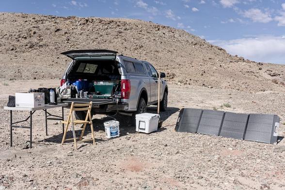 A color landscape photo showing a car camping setup on a barren dirt pad with a hill in the background. A grey pickup truck with a shell on the pickup bed is facing away from the camera and toward the right. The back is open. A table extends out the back on the right and has various items on it. A green two burner Coleman stove is on the right side of the open tailgate. A wood chair is in the corner formed by the table and tailgate. A white ice chest sets to by the right read tire. A a four section solar panel extends out from the right side of the truck.