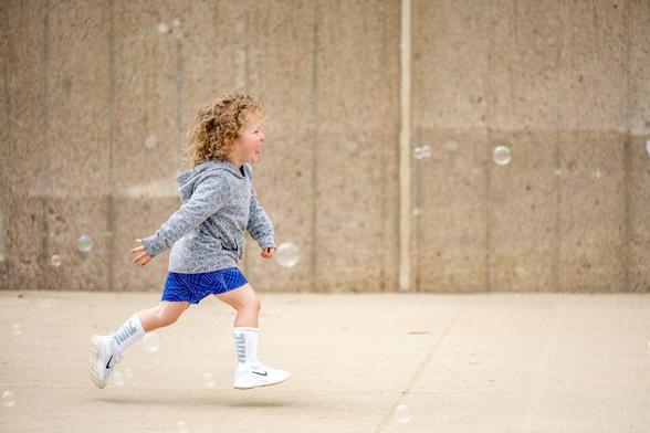A young boy with curly blonde hair, wearing a gray hooded sweatshirt, blue athletic shorts, white tube socks, and white sneakers, chases after bubbles in the air. The photo is taken as a profile from his right side. His face shows pure joy as he tries to capture and pop the bubbles.