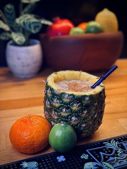 cocktail with grated nutmeg garnish in a hollowed-out pineapple with a blue surfside sips glass straw sitting next to citrus with a fruit bowl in the background