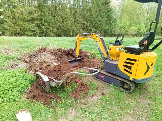 A yellow and black mini excavator with a wide, toothless grading bucket has turned over a large compost pile near a forest. Fresh compost is exposed and dumped into a wheelbarrow standing aside, somewhat spilling over.