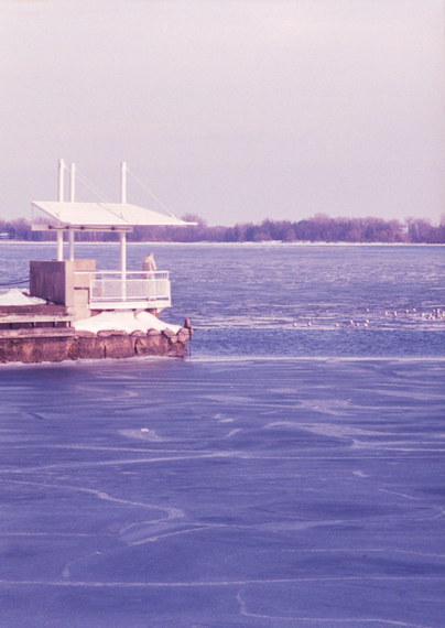 A person in a puffy, white winter coat stands on a covered observation deck at the end of a concrete pier, the water is almost entirely covered by a thin layer of ice on which gulls have gathered.