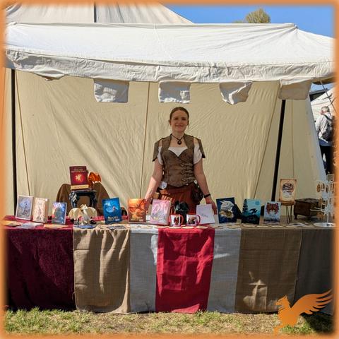Foto von mir (Frau Ende 20 in Mittelalter- bzw Fantasyoutfit) in einem Mittelalterzelt. Auf einem langen Tisch mit rotbraunen Decken sind Fantasybücher, Tassen und hölzerne Schlüsselanhänger ausgestellt.