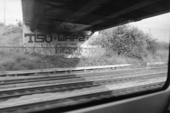 A black and white photograph taken through a train window at the underside of a bridge containing graffiti.