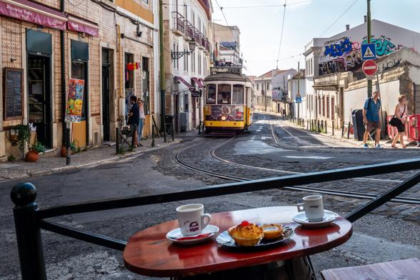 Straßenszene in Lissabon mit vorbeifahrender gelber Straßenbahn der Linie 28. Im Vordergrund ein Café-Tisch mit Espresso und Pastéis de Nata, im Hintergrund Häuser mit Fliesenfassaden, Passanten und Graffiti.
