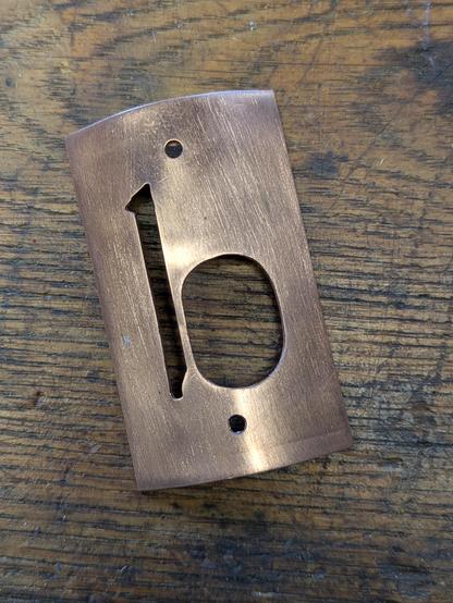 Das Bild zeigt ein metallisches Etikett mit einem bronzefarbenen Finish, das auf einem Holztisch liegt. Das Etikett hat eine rechteckige Form mit abgerundeten Ecken und zwei Löcher oben, die für Schrauben oder Nägel verwendet werden können. In der Mitte des Etiketts ist eine numerische Form "10" ausgeschnitten, wobei die "1" vertikal und die "0" horizontal angeordnet ist. Die Oberfläche des Metalls hat eine matte, gebürstete Textur, die ein rustikales Aussehen verleiht. Der Holztisch im Hintergrund hat eine dunkle, verwitterte Oberfläche mit sichtbaren Holzfasern und Kratzern, was auf eine natürliche und robuste Umgebung hinweist.
