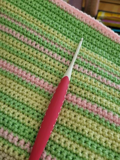 Das Bild zeigt ein gehäkeltes Stück Stoff mit horizontalen Streifen in abwechselndem Hellgrün, Zartgelb und sanftem Rosa. Die Textur des Häkelmusters ist deutlich erkennbar. Quer über das Häkelstück liegt eine Häkelnadel. Sie hat einen glatten, pinkfarbenen Griff, der sich zum weißen, leicht glänzenden Haken hin verjüngt. Die Nadel liegt diagonal auf dem Stoff und deutet auf ein Handarbeitsprojekt hin.