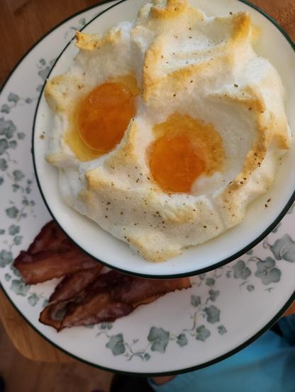 A white bowl sits in a while plate. The plate has several slices of bacon. The bowl is full of lightly toasted egg white meringue, with two egg yolks baked into the top.
