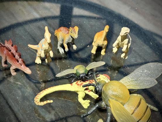 A green Dilophosaurus with brown spots is on their side while two yellow bees, one large, the other small, attack them. A cult of dinosaurs watches on. From left to right: A red Stegosaurus, a yellow Parasaurolophus with blue stripes, a brown and grey Dilophosaurus, an orange and yellow Brachiosaurus, and a white Pachycephalosaurus. All stand on a circular, glass table. Bits of green and yellow pollen dot the surface of the table.