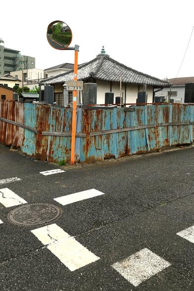 A rusty fence made from blue corrugated metal that has gotten out of shape in many places separates a wet intersection with bright white road markings in the foreground from the cemetery and temple behind said fence. The temple is nothing special, single-storey from old weathered wood with the characteristic slanted roof. It's the contrast in colors, the wet asphalt, the wonky rusty fence, and the final resting places around the temple that caught my attention. It didn't hurt that a traffic mirror on an orange pole also made the shot.