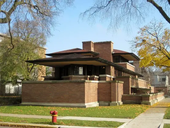 This image shows a distinctive architectural structure known for its horizontal lines and integration with the landscape. It features a combination of brick and large overhanging roofs, typical of the Prairie School architectural style. The building is surrounded by trees and grassy areas, indicating it is situated in a suburban environment. The design emphasizes open spaces and connection to nature.

Image Credits: Wikimedia / Teemu08 / CC BY-SA 3.0
