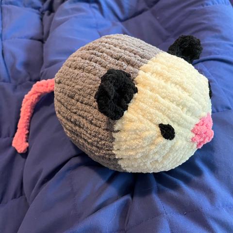 A large knitted opossum made of fuzzy chenille yarn, maybe 8 inches across, with a blunt pink nose and embroidered black eyes