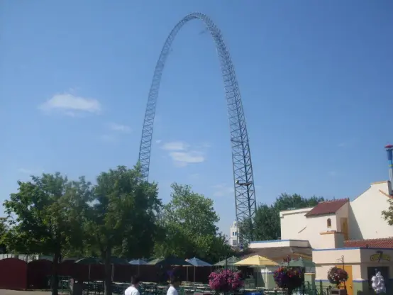 The image shows a large, arch-shaped structure that appears to be an amusement park ride, likely a roller coaster or a similar attraction. There are trees and some buildings in the vicinity, indicating an outdoor setting, possibly within a theme park. The sky is clear and blue, suggesting a sunny day.

Image Credits: Wikimedia / Katarighe. / CC BY-SA 3.0