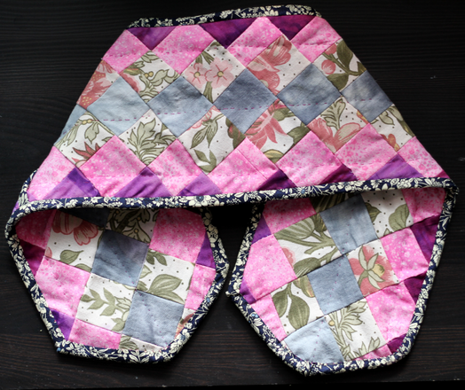 A pair of oven mitts that are conjoined into one long strip. The mitts are multi coloured with a lot of pink
