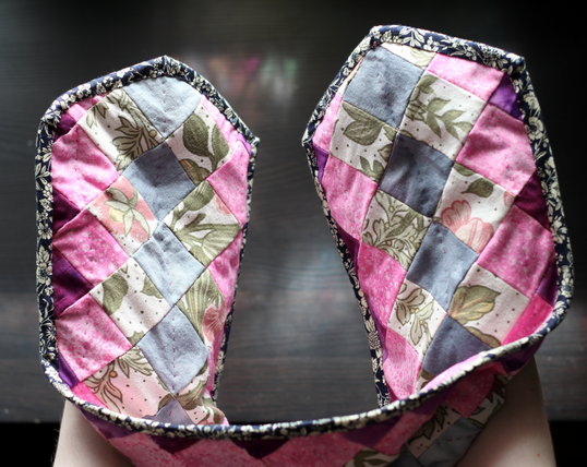 A pair of oven mitts that are conjoined into one long strip. The mitts are multi coloured with a lot of pink