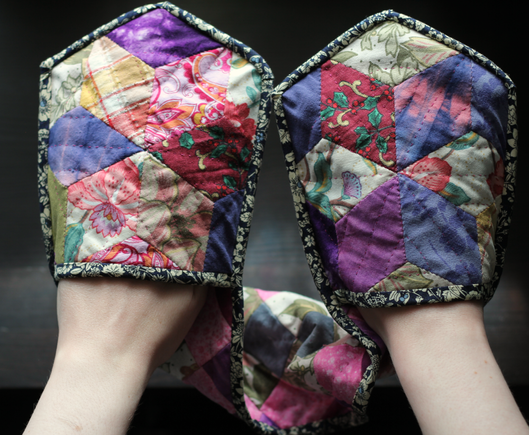 A pair of oven mitts that are conjoined into one long strip. The mitts are multi coloured with a lot of pink