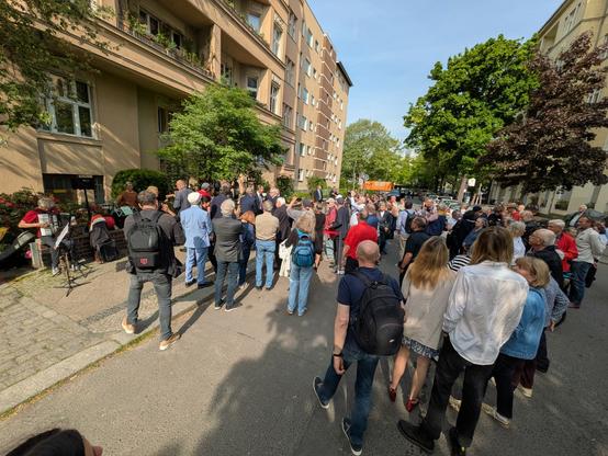 Eine kleine Menschenmenge auf einer ruhigen, kleinen Straße vor einem großen, hellbraunen Altbau-Haus. Die meisten der Menschen sind von hinten zu sehen, viele von ihnen fotografieren.