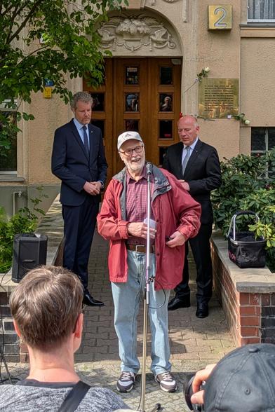 Ein älterer Mann mit weißem Vollbart und Brille steht an einem Mikrofon, das auf einem Ständer befestigt ist. Der Mann trägt eine rote Jacke, ein kariertes Hemd, hellblaue Jeans und Sportschuhe, dazu eine hellgraue Schirmmütze auf dem Kopf. Er lächelt und zieht einen Zettel aus seiner Jackentasche und scheint ins Mikrofon zu sprechen. Im Hintergrund sieht man einen Hauseingang mit einer Messingtafel und der Hausnummer 2 darüber. Vor dem Hauseingang stehen zwei Männer, beide tragen dunkelblauen Anzug und Krawatte. Der Mann links ist sehr groß und hat lockige, graue Haare. Der Mann rechts ist deutlich kleiner und hat eine markante Glatze – es ist Kai Wegner, der Regierende Bürgermeister von Berlin.