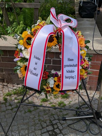 Ein Blumenkranz. Er besteht hauptsächlich aus weißen und gelben Blumen, u.a. erkennt man Sonnenblumen und weiße Rosen. Um den Kranz ist eine weiße Schleife mit roten Rändern gebunden. Die Schleife trägt die Aufschrift "Frieden ist der Ernstfall" und "Hausgemeinschaft Schulenburgring 2".