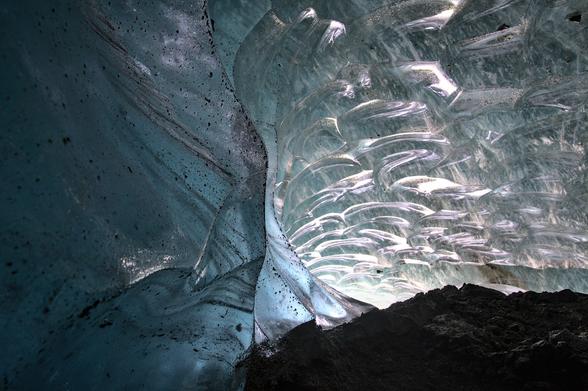 View from inside a glacier cave beneath the Canwell Glacier in Alaska. The foreground shows smooth, bluish ice walls dusted with dark debris. Light filters in from a translucent, textured ceiling where sculpted ice arcs reflect and scatter light, resembling frozen ripples. A ridge of dark rocky ground runs along the base of the cave.