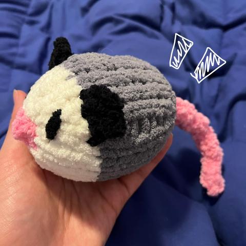 A hand holding a small knitted opossum made from fuzzy chenille yarn. He has embroidered black eyes, a little pink nose, black ears, and a pink knitted cord for a tail. He's maybe 3 or 4 inches wide