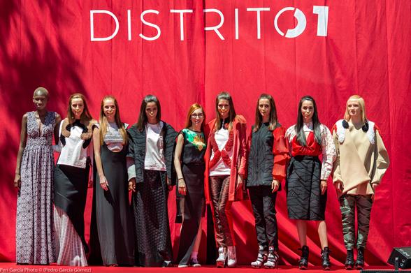 Foto grupal de ocho modelos y la diseñadora Leyre Valiente en el desfile de moda urbana Distrito 1. Domina la escena el telón rojo con ese nombre escrito delante de ellas.