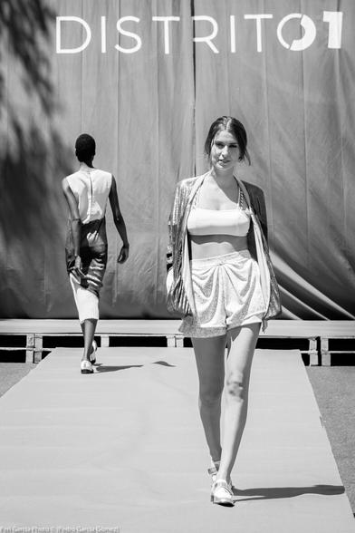 Foto en blanco y negro y plano entero frontal y en vertical de una de las modelos que avanza hacía el frente por la pasarela mientras otra avanza hacía el fondo (el telón rojo con el texto Distrito 1 impreso). 

Va vestida con un pantalón corto y un top, ambos de color blanco y una chaquetilla abierta de color gris claro. Es de piel muy blanca y pelo castaño y es de cuerpo exuberante.
