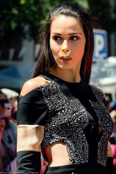 Foto en color, plano medio frontal y vertical de una de las modelos que mira a la izquierda con el fondo desenfocado del público, carpas del festival y los edificios del fondo. Destaca que la modelo lleva un gran aro en la nariz en forma de media luna de color dorado.

Es de piel blanca rosada y de pelo y ojos negros. Lleva un vestido de color negro con parte del pecho con un motivo rayado blanco y negro y una sobremanga beige. Con los hombros y la cintura descubiertos.