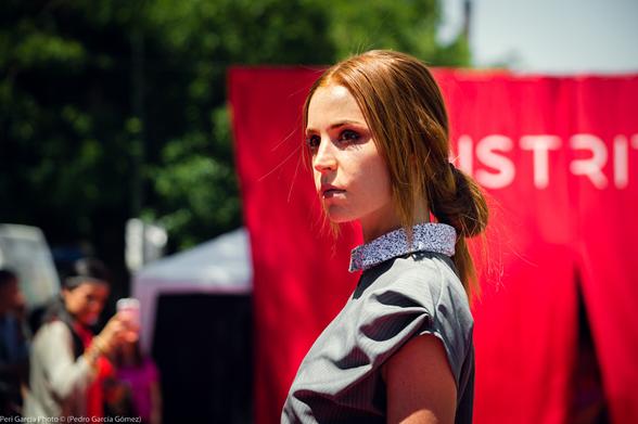 Foto en color y primer plano semifrontal de perfil una de las modelos mirando a cámara  a la izquierda con el fondo desenfocado de copas verdes de los arboles, público y el telón. La modelo es de piel blanca y pelirroja con el pelo recogido en un coleta baja. Va vestida con un vestido sin mangas y de color gris con el cuello de colores blanco punteado en negro.