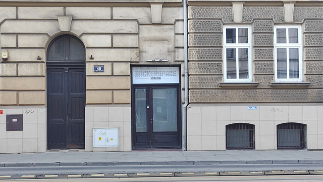 A photo of the entrance to Hackerspace Kraków at 46 Limanowskiego Street. The door is glass with a mesh pattern, framed by dark wood, and above it is a sign reading "HACKERSPACE KRAKÓW" in bold, geometric letters. The building has a classical European facade with stone detailing, large windows, and tram tracks visible in the foreground.