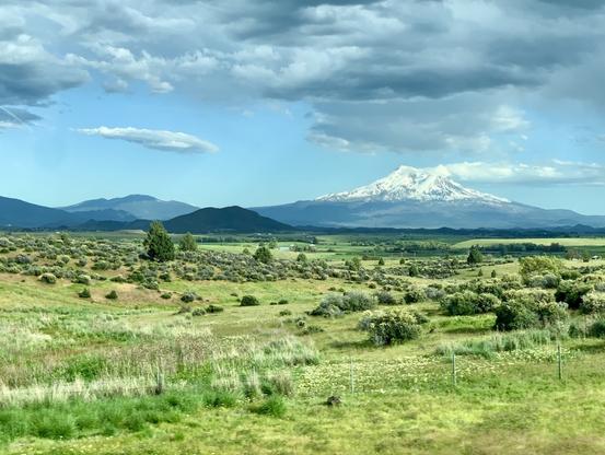 The shows a snow capped stratovolcano on the horizon near the upper right third. Cumulus clouds hang over it and other mountains along the horizon. The bottom half of the picture is a mix of chaparral and grassy fields.