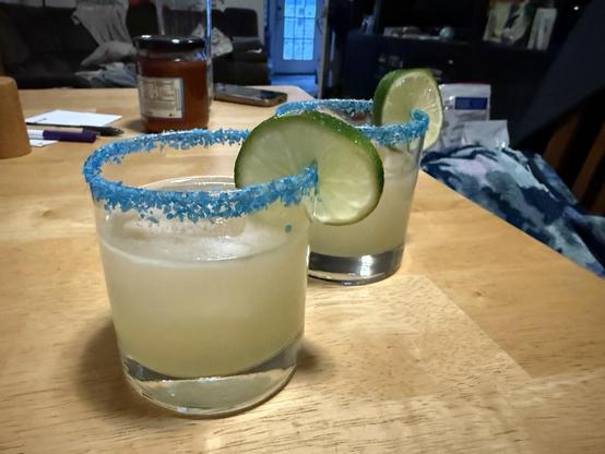 Two rocks glasses with a lime colored cocktail garnished with blue salt and a slice of lime.