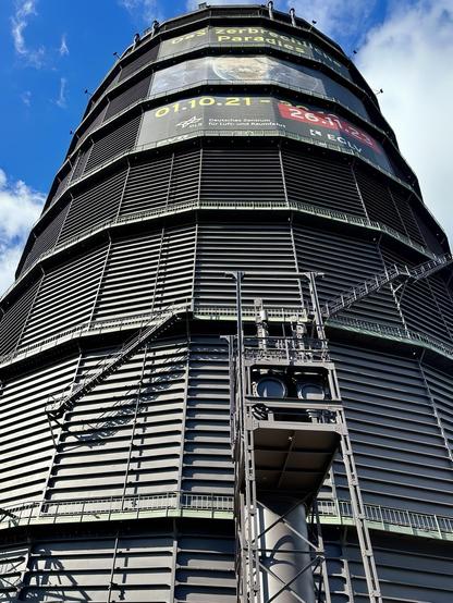 This image shows a towering cylindrical structure, possibly a modern observation tower or a part of an industrial facility. It features several digital displays displaying information like dates and some names, as well as various digital and analog clocks. The structure is clad in metal with horizontal louvers, and there are external staircases winding around it. There are also logos and text that indicate the involvement of "Deutsches Zentrum für Luft- und Raumfahrt," which is the German Aerospace Center, suggesting that this structure might be related to aerospace or scientific research.