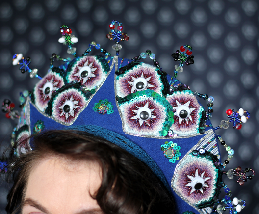 A blue crescent shaped headdress on a woman’s head against a blue background. The headdress is decorated with sequins and mirrors and beads in a pattern that looks like peacock feathers.